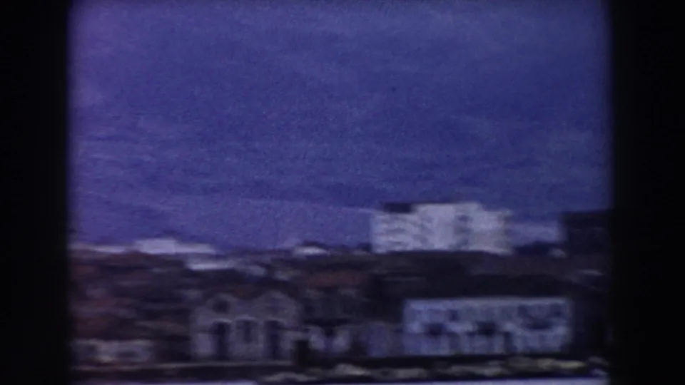 1959 home-movie view near Pisa showing rooftops, a pale building, and dark hillside terrain in soft focus.