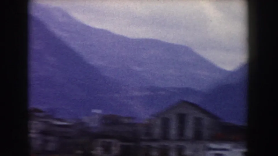 Dim 8mm frame near Pisa with a mountain slope rising behind a small foreground building and clustered dark structures.
