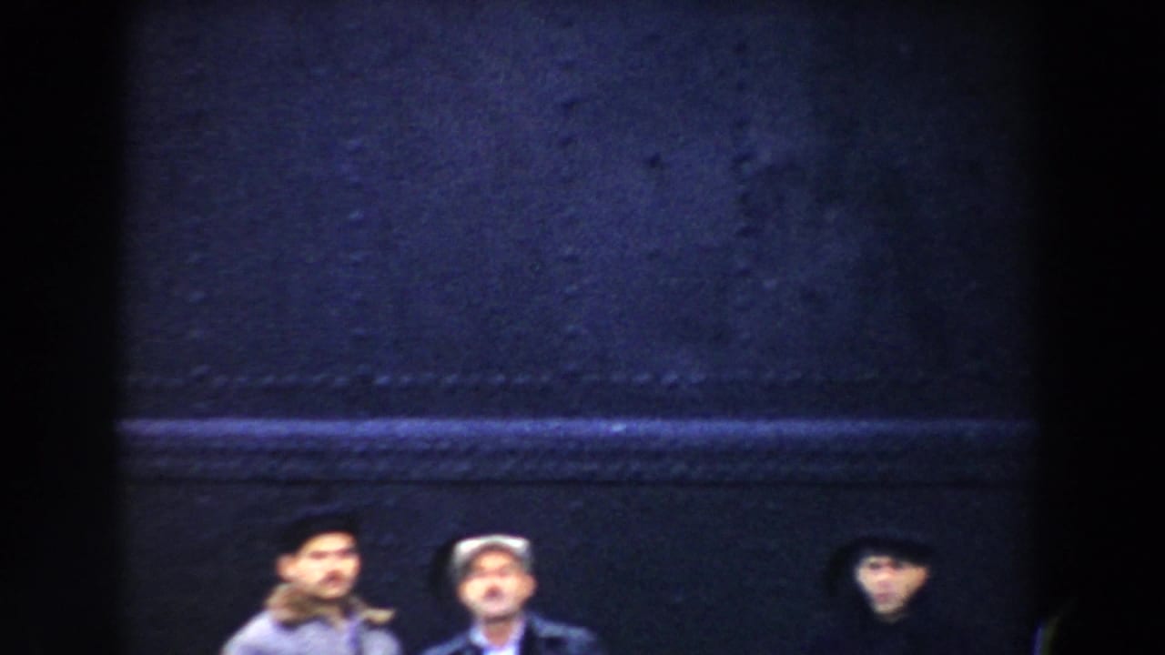 Small figures stand below a dark dockside or ship structure in a grainy frame.