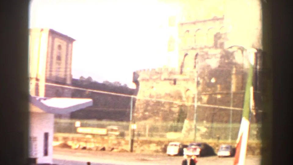Overexposed 1959 Pisa travel frame with historic stone walls, parked cars, nearby buildings, and an Italian flag at the edge.