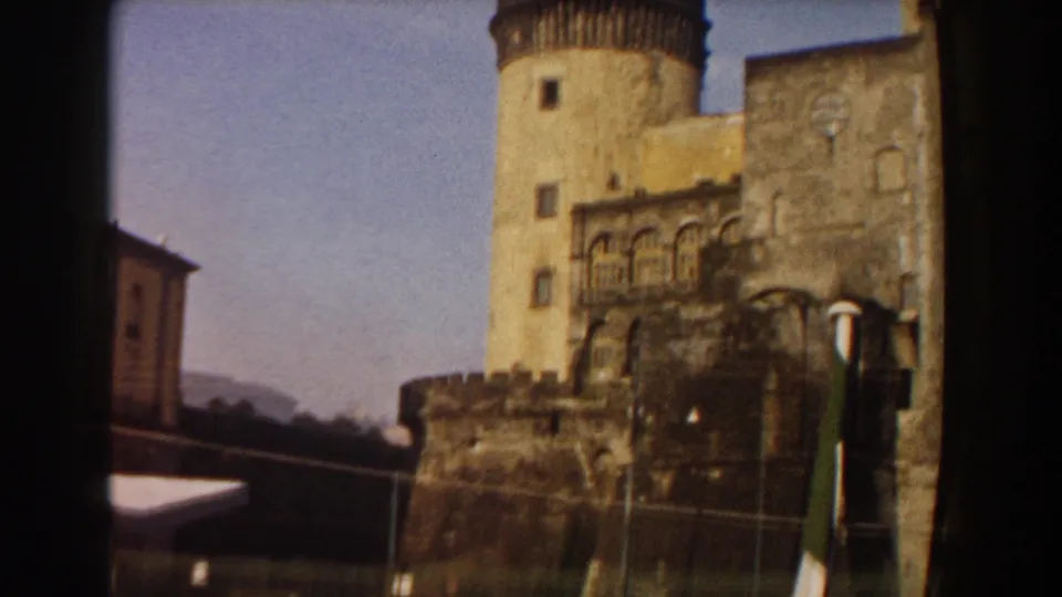1959 Pisa home-movie view of a towered stone waterfront structure with crenellated walls, parked cars, and an Italian flag.
