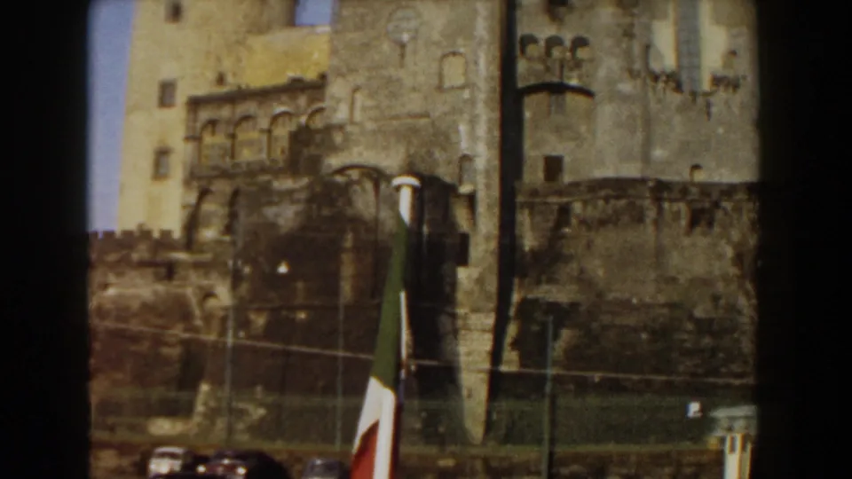 1959 8mm street-level view in Pisa shows the castle wall, a flagpole, lamps, fencing, and parked cars near the landmark.