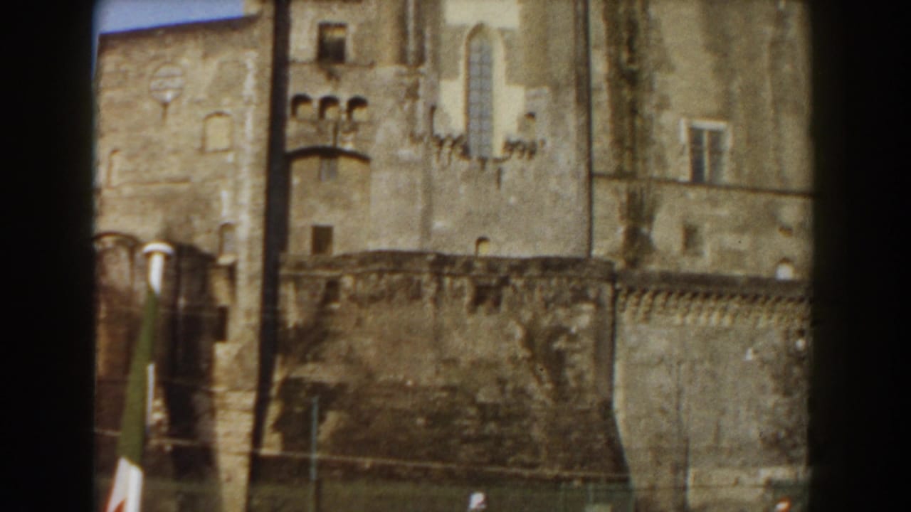 1959 Pisa home-movie frame showing a weathered stone castle exterior with battlements, arched openings, and a partial flag in front.