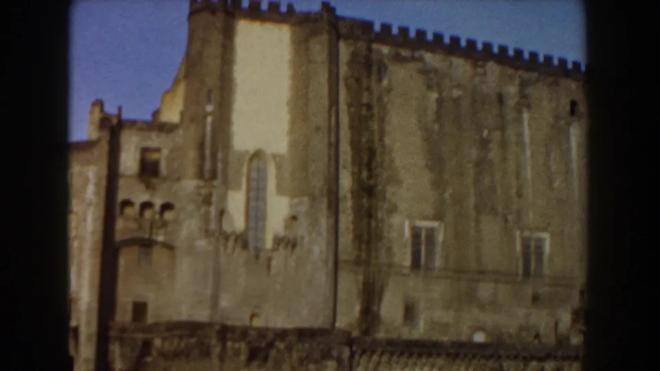 1959 Pisa frame of a historic castle exterior, with aged stone surfaces, small windows, crenellations, and clear blue sky.