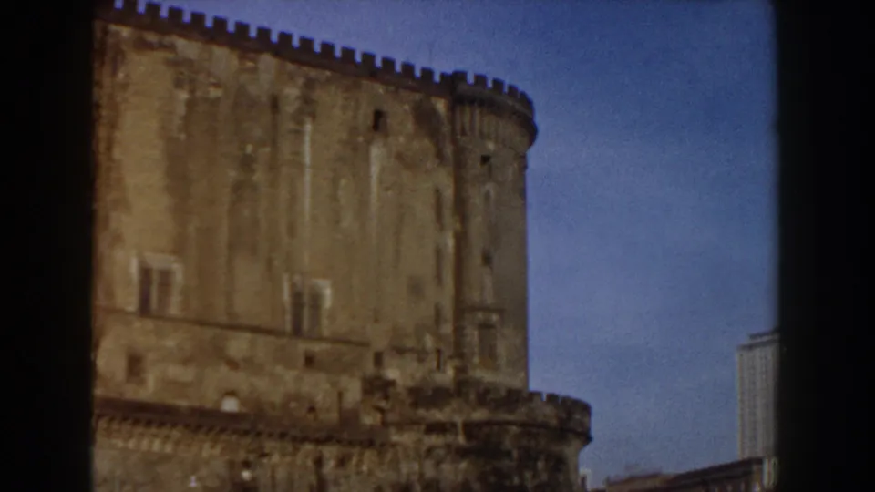 1959 Pisa frame showing a weathered castle tower and wall with small windows, battlements, blue sky, and a building edge at right.