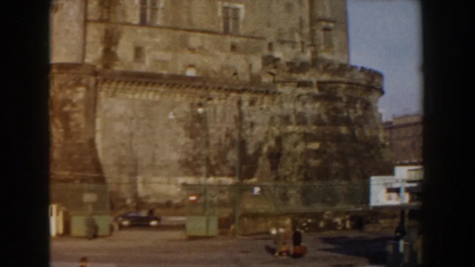 Pisa, Italy travel footage frame showing the base of a weathered fortress wall with cars, street fixtures and small pedestrians below.