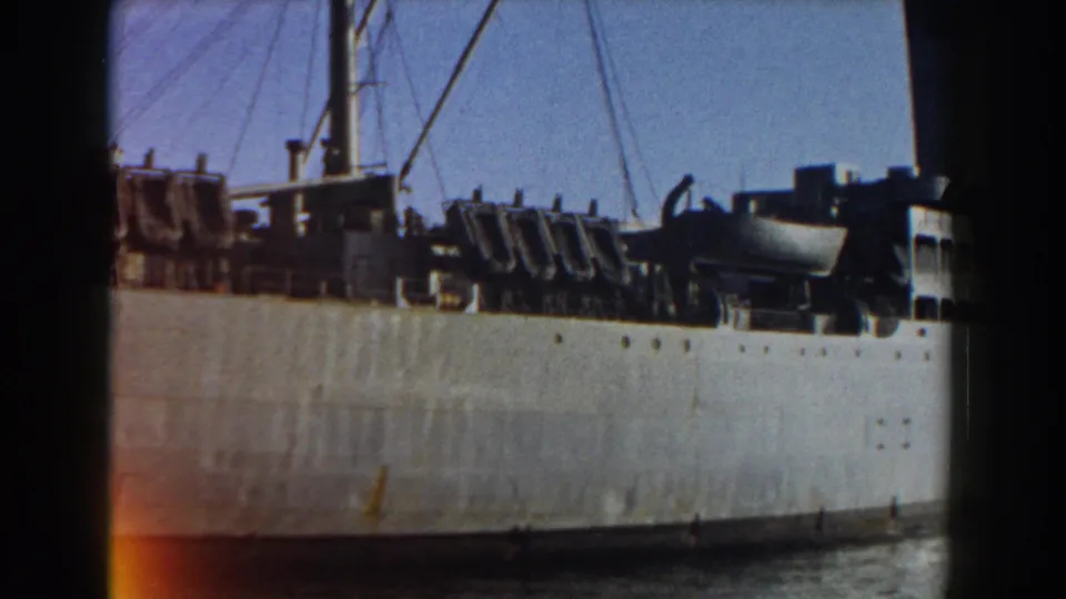 Large cargo ship docked in Pisa, Italy, with pale hull, dark deck structures, rigging, and water at the base.