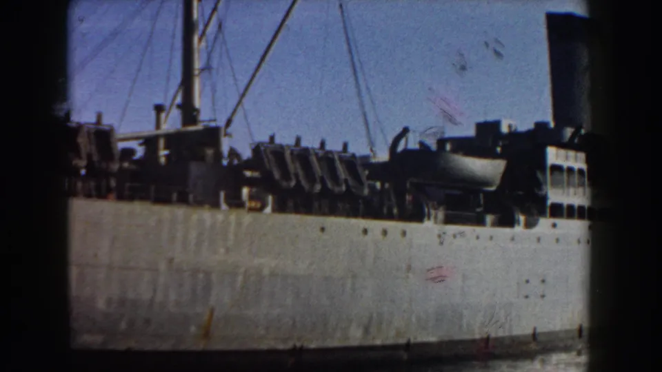 1959 8mm view of the docked cargo ship's side, showing its pale hull, deck gear, mast lines, and waterline.
