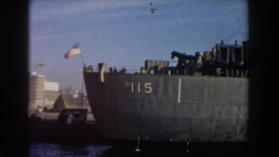 A closer Pisa harbor frame shows the P115 marking on the ship hull, the American flag, faint deck silhouettes, dockside vehicles, and blue sky.