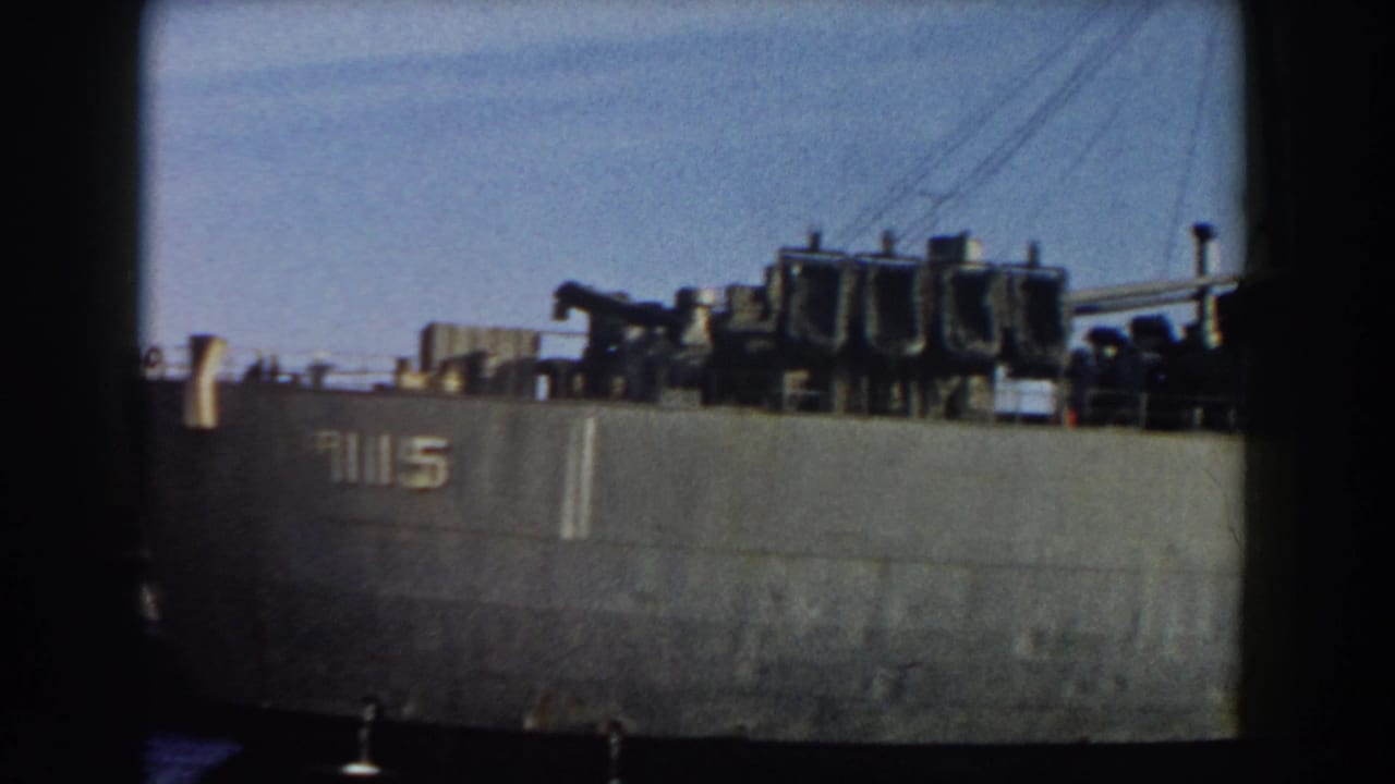 Gray military ship hull with visible numbers and dark deck equipment under a blue sky.