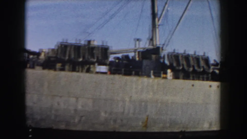The camera pans along the 1959 Italy harbor view, showing deck machinery and rigging on a moored military ship.
