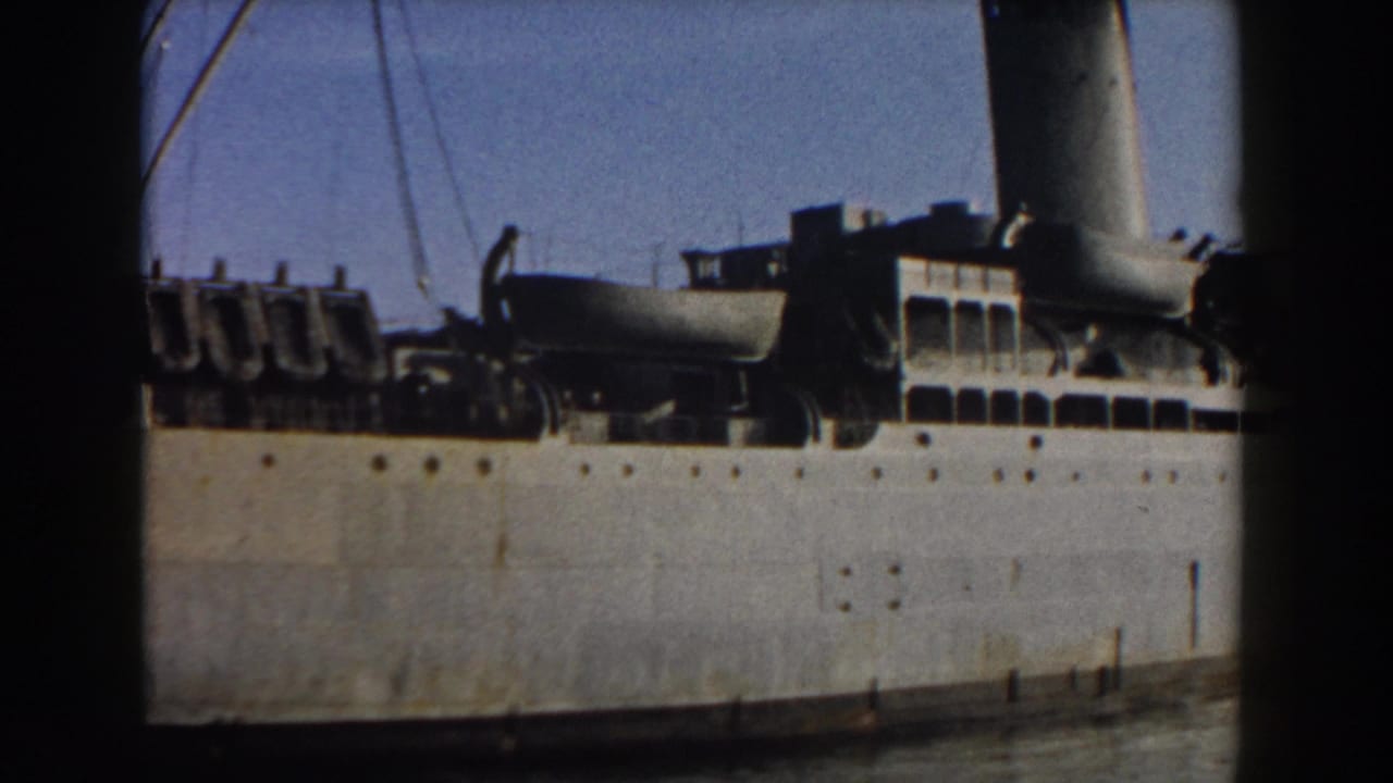 Large white ship hull with portholes, deck structures, and a tall funnel at port.