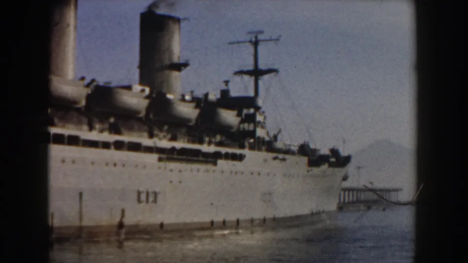 1959 Pisa, Italy frame of a stationary passenger ship beside calm water, with smokestacks, mast rigging, and hazy mountains beyond.