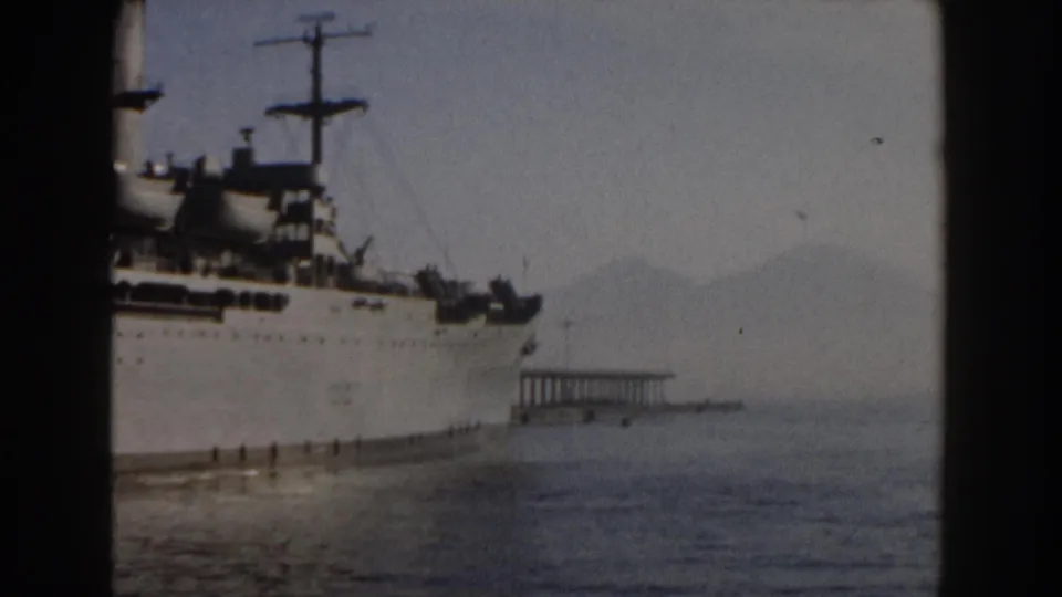 1959 Pisa, Italy frame showing the stationary white ship at harbor, framed by dark film edges with water and mountains behind it.