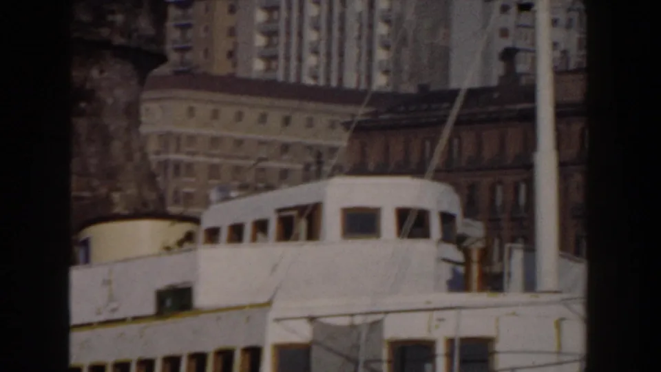 1959 Pisa home-movie frame showing a white boat or ferry structure before mixed waterfront architecture and tall background buildings.