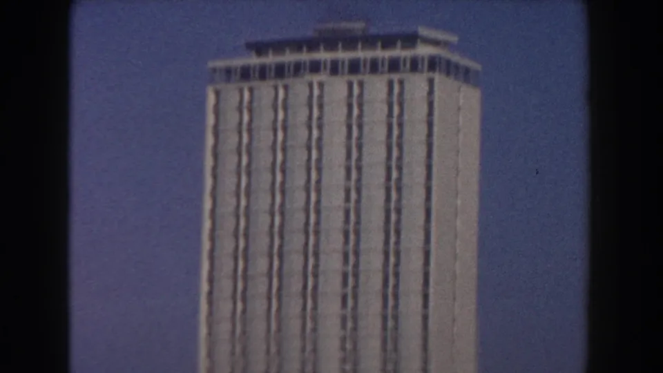 1959 Pisa frame centered on a tall modern-looking building with vertical window bands, seen through soft 8mm motion blur.