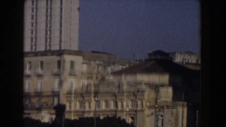 Late-frame 1959 Pisa view with older low-rise facades, the edge of a taller tower, rooflines, and dark foreground framing from the moving camera.