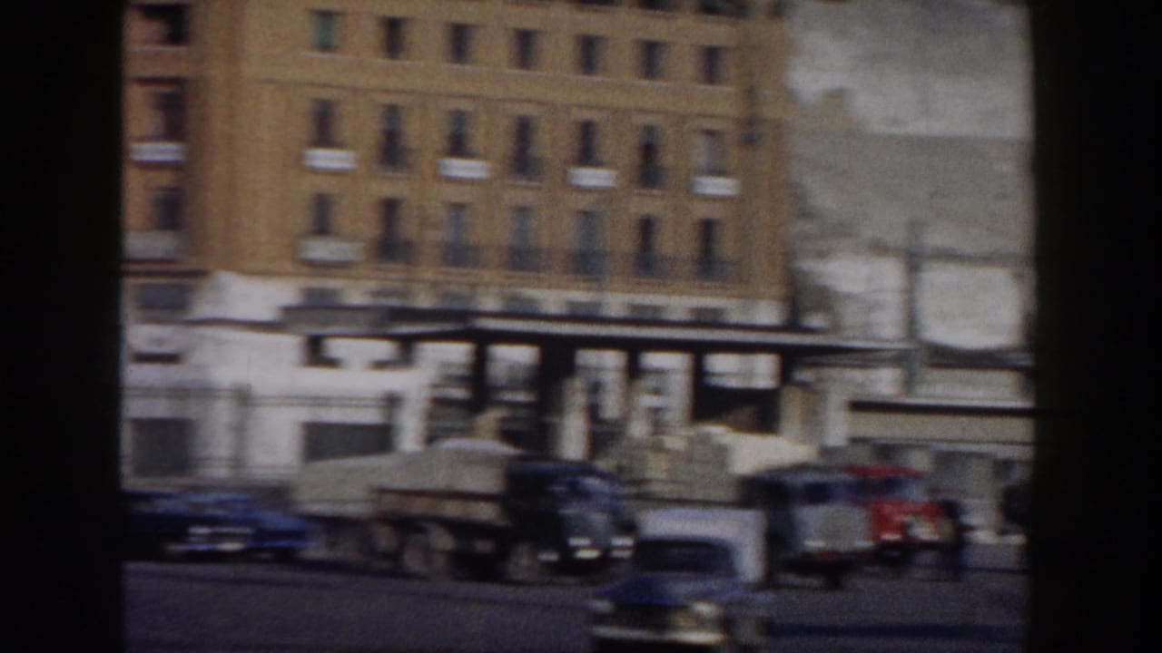 Urban street with parked cars, delivery trucks and multi-story buildings behind storefronts.
