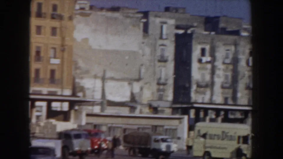 1959 Pisa street scene with parked trucks and cars in front of weathered buildings and low shopfronts.