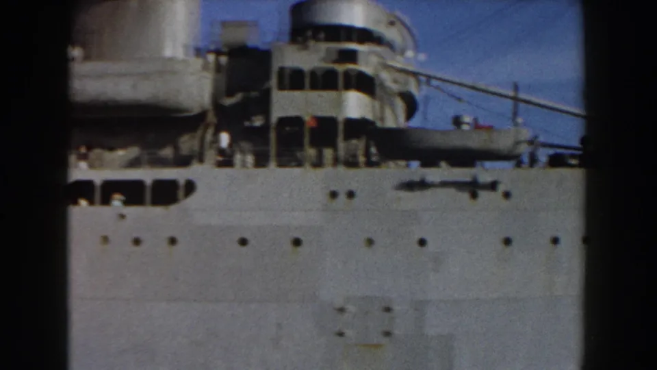 The marked naval vessel continues past the camera, showing its upper decks, funnel, railings, and portholes against clear sky.
