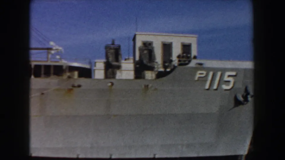 1959 Pisa, Italy frame showing the grey ship's bow with hull number P115 beside waterfront buildings and blue sky.