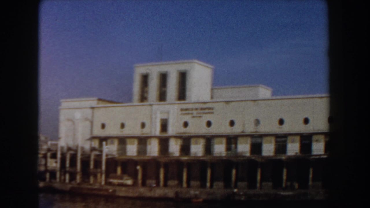 White waterfront building with circular windows, signage, pier supports, dark water, and blue sky.