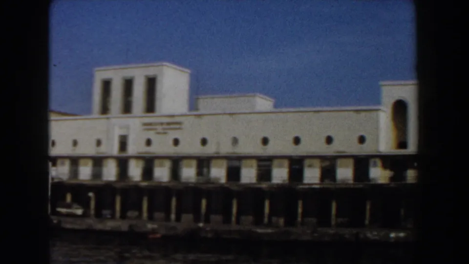 Passing 8mm view of a long white Pisa waterfront facade with circular windows, a raised central section, pier supports, and water below.