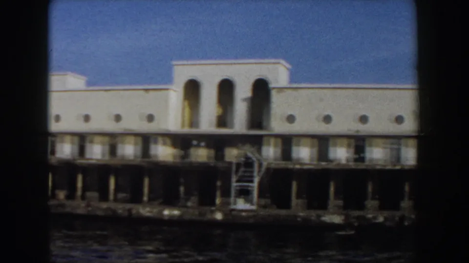 1959 Pisa waterfront building detail showing tall arched openings, round windows, exterior stairs, pier columns, and water in the foreground.