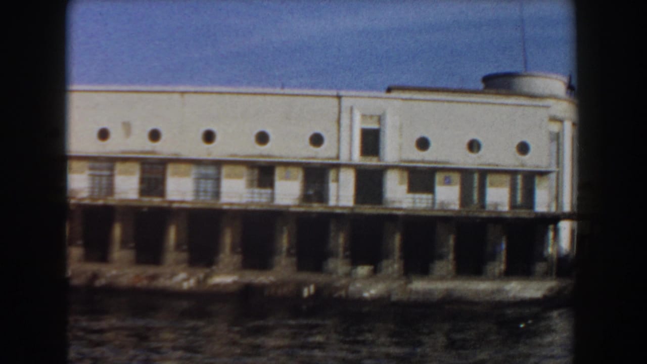 White waterfront building on pilings beside calm water, with circular windows and dark pier openings.