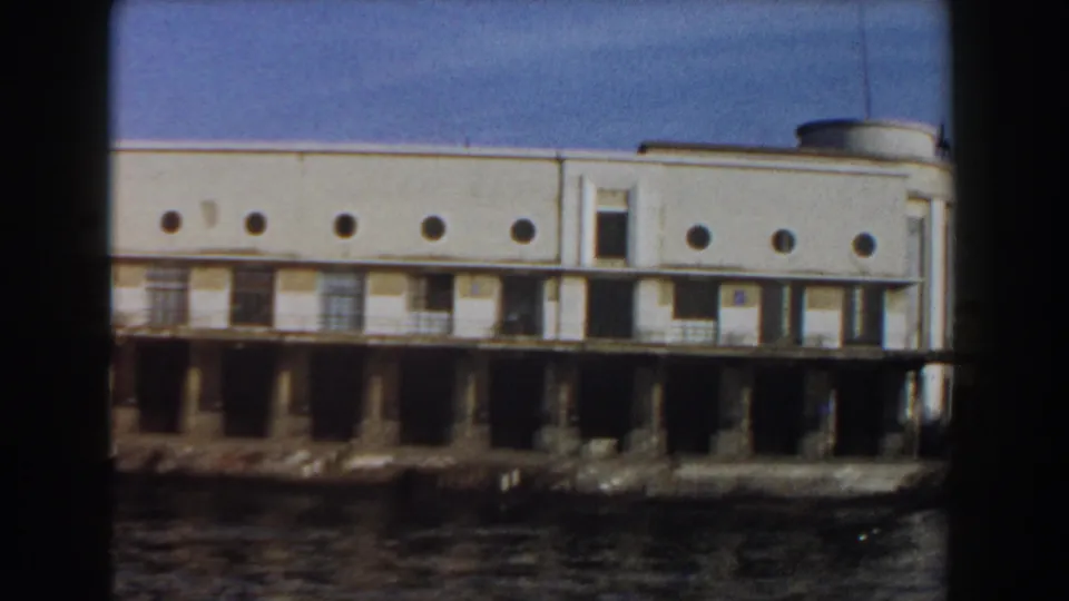 1959 Pisa, Italy waterfront view showing a white pier building with circular windows, weathered pilings, and water in the foreground.