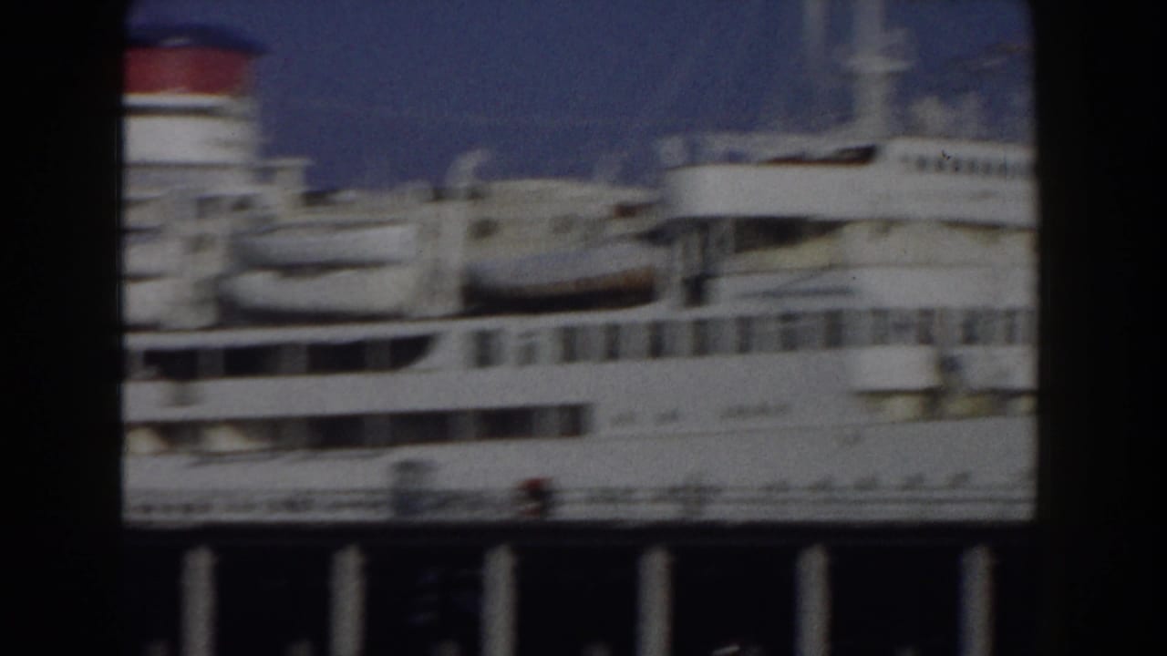 1959 8mm Italy travel footage showing a large white passenger ship moored near the coast, partly framed by dark harbor structure.