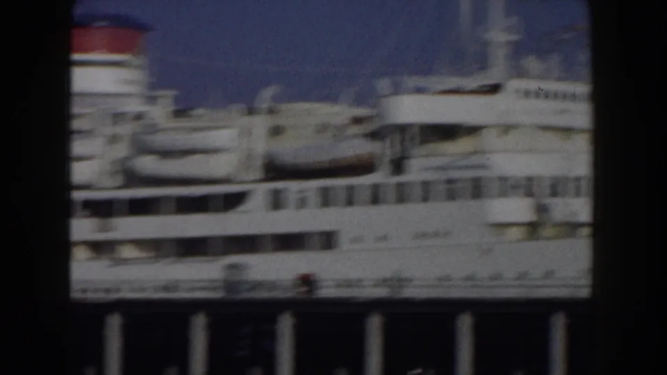 1959 8mm Italy travel footage showing a large white passenger ship moored near the coast, partly framed by dark harbor structure.