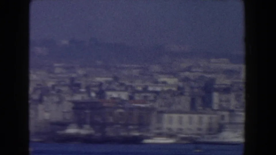 1959 8mm view across water toward a hazy Italian waterfront cityscape, with low buildings and small boats along the shore.