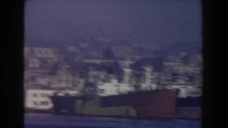 1959 8mm harbor view near Pisa, showing moored vessels, shoreline buildings, and hazy hills beyond the coastal city.