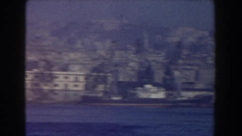 1959 8mm Italy harbor footage showing a moored vessel and waterfront buildings across blue water, softened by haze and film grain.