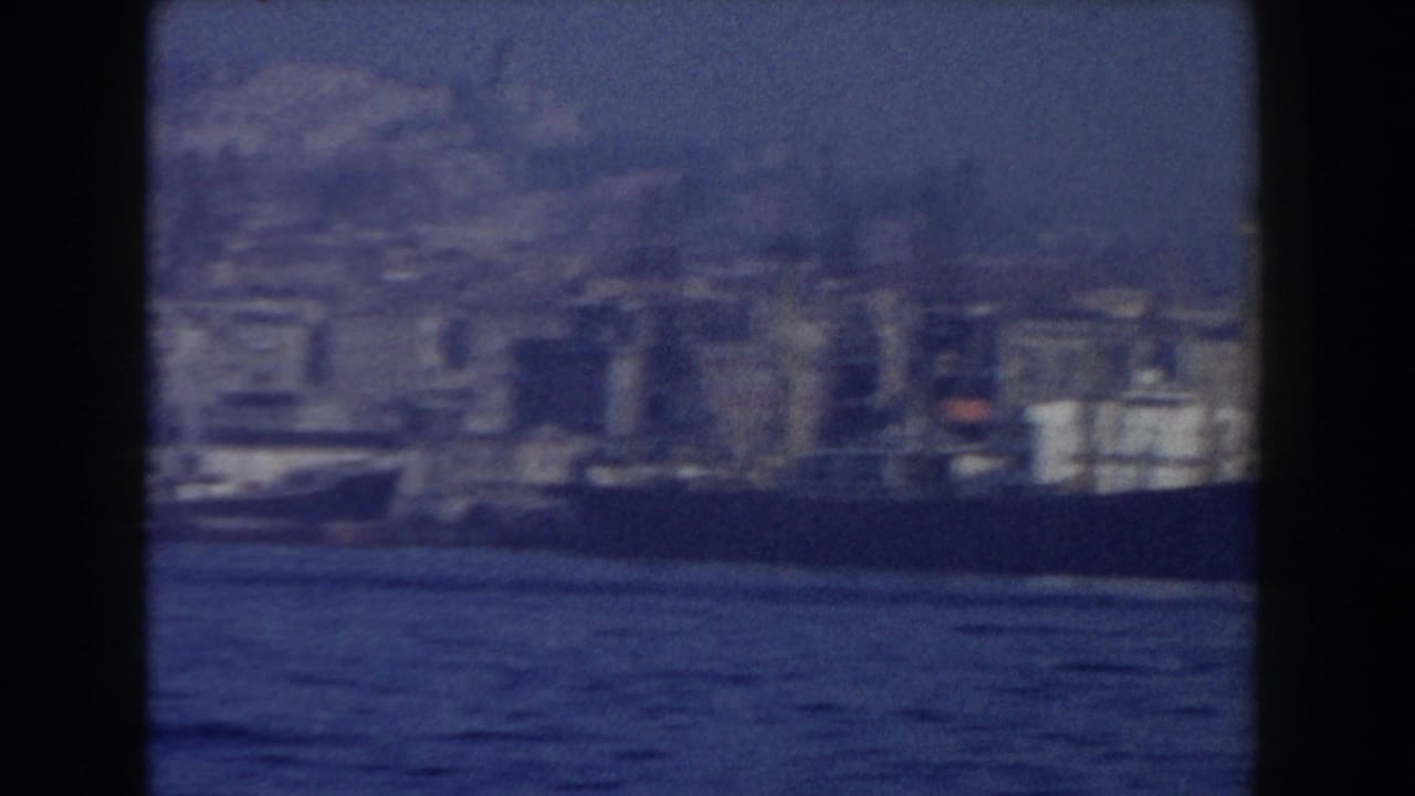 1959 8mm view from a boat toward a hazy Pisa waterfront with water in the foreground and low buildings along shore.