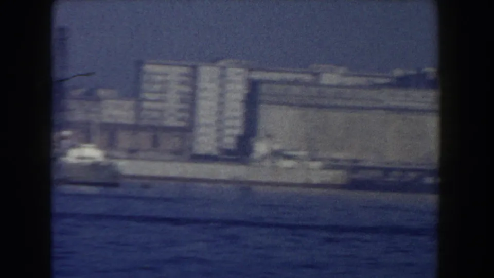 1959 boat-shot view of low waterfront buildings in Pisa, with blurred vehicles or light shapes near the shore.