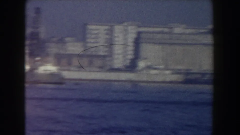 A soft 8mm frame shows Pisa waterfront buildings across the water, including larger rectangular structures and a dark scratch mark.