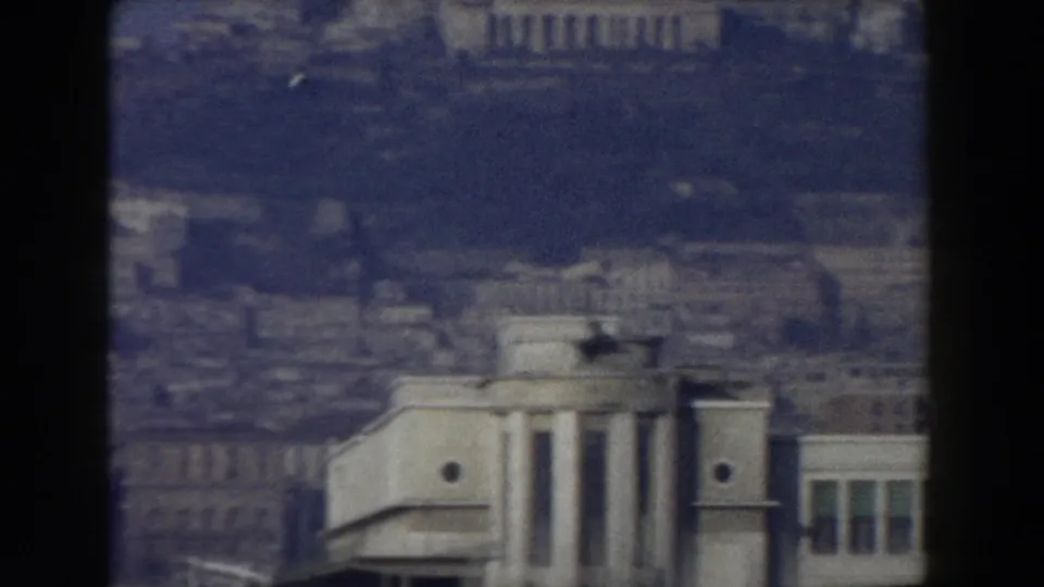 Distant 1959 Pisa view with a pale classical-looking facade, clustered buildings, haze, and visible film-frame edges.