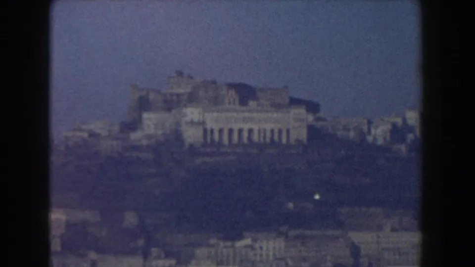 1959 Pisa travel-film frame showing a distant historic structure on higher ground, with rooftops below and a blue hazy sky.