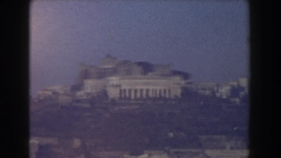 1959 Pisa home-movie frame showing distant pale landmark buildings through haze beneath a blue sky.