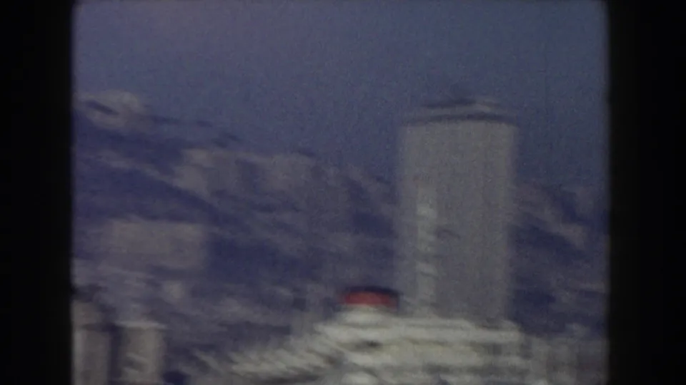 1959 Pisa home-movie frame showing a tall cylindrical structure, rooftops, and hazy distant buildings.