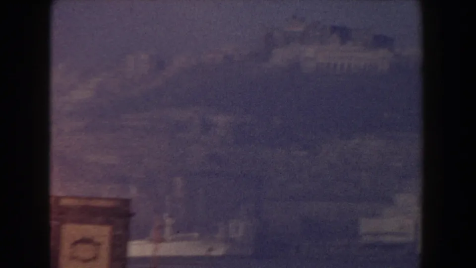 Blue-tinted 8mm frame showing faint Pisa rooftops and hillside structures through heavy haze.