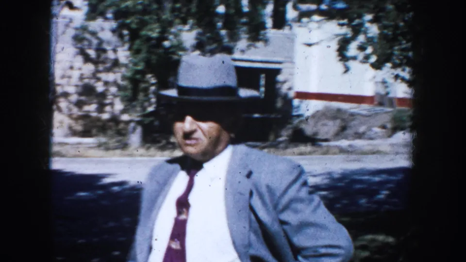 Adult man in a light gray suit, dark tie, and fedora walks through a sunlit South Dakota residential setting in 1953.