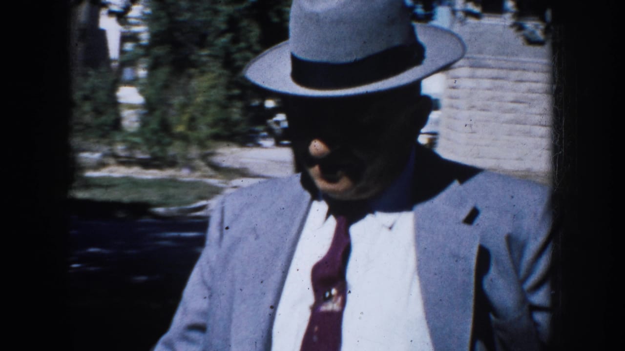 Closer 8mm frame of the adult man in fedora, suit, white shirt, and tie moving past the camera outdoors.