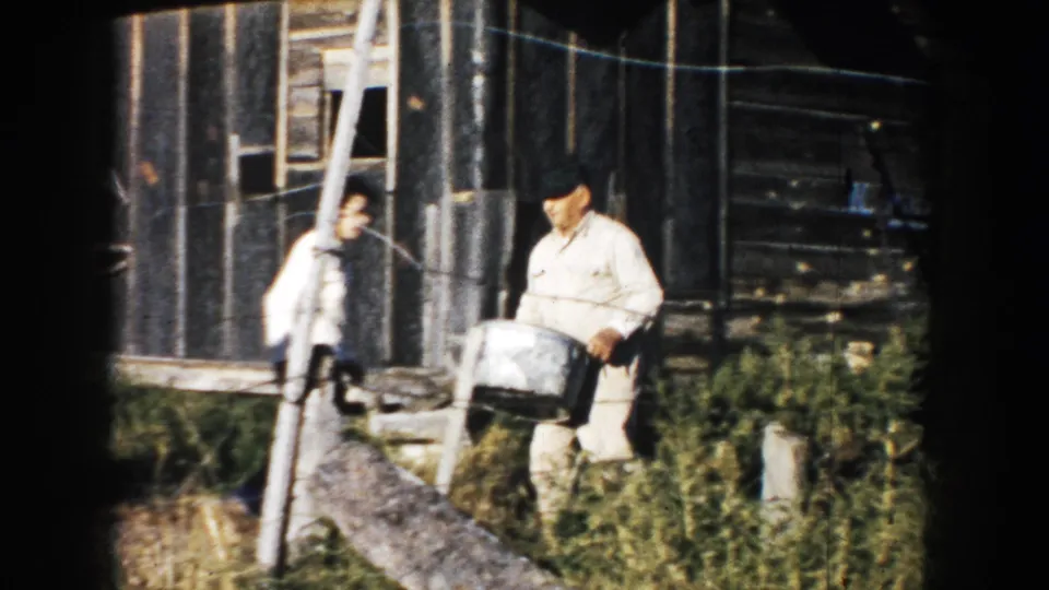 Two adults in light shirts stand outside a weathered barn, with one carrying a metal container in 1953 South Dakota footage.