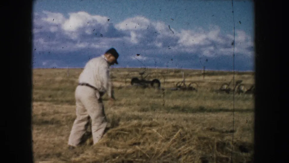 A man in a light shirt walks across a dry South Dakota field under a cloudy sky, with distant farm equipment on the prairie.