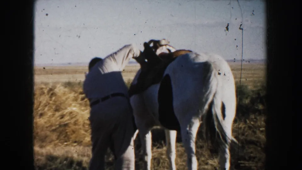 1953 South Dakota frame of a man working close to a white horse, appearing to adjust saddle or bridle tack in a farm field.