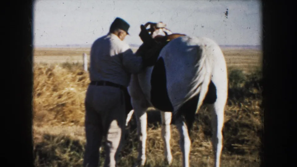 A man in a dark hat stands close to a black-and-white horse, adjusting saddle gear in a South Dakota prairie setting.