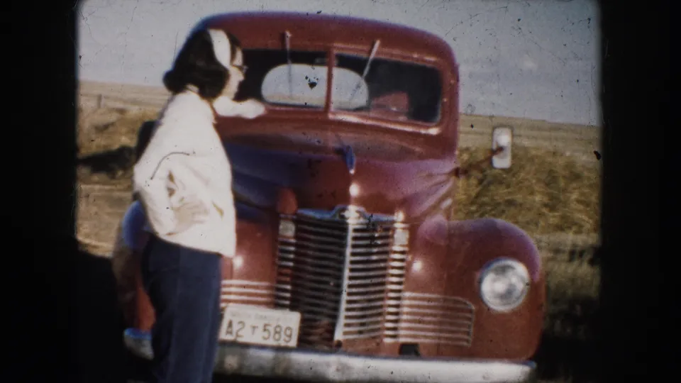 Woman in a light jacket and dark trousers stands near a red truck in a South Dakota field in 1953 footage.